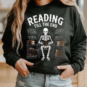 May include: Dark gray sweatshirt with the text "READING TILL THE END" above an illustration of a skeleton reading a book while seated in a chair. Bookshelves and stacks of books surround the skeleton.