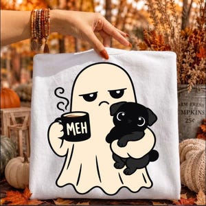Könnte beinhalten: Ein weißes T-Shirt mit einer Cartoon-Grafik eines cremefarbenen Geistes mit gelangweilt-mürrischem Gesichtsausdruck. Der Geist hält eine schwarze Tasse mit der weißen Aufschrift MEH und trägt einen kleinen schwarzen Welpen im Arm.