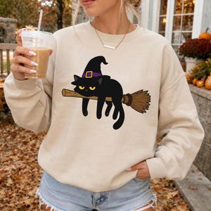 May include: A beige crewneck sweatshirt featuring a graphic of a black cat wearing a purple-banded witch hat, lying across a brown broomstick. The cat has yellow eyes and is positioned centrally on the front of the long-sleeved pullover garment.