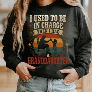 Może przedstawiać: Ciemnoszara bluza z napisem "I USED TO BE IN CHARGE THEN I HAD A GRANDDAUGHTER". Grafika przedstawia dziadka i wnuczkę dających sobie piątkę na tle zachodu słońca.