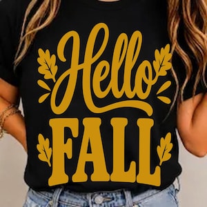 Może przedstawiać: Czarny t-shirt z krótkim rękawem z napisem Hello FALL wykonanym pogrubioną czcionką w kolorze musztardowym. Wzór jest ozdobiony stylizowanymi ilustracjami liści dębu w kolorze musztardowym, umieszczonymi wokół tekstu na przodzie odzieży.