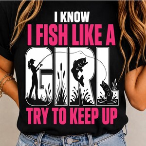 May include: A black t-shirt featuring the white and pink text I KNOW I FISH LIKE A GIRL TRY TO KEEP UP. The design includes a silhouette of a person fishing, cattails, and two jumping fish integrated into the large white letters of the word GIRL.