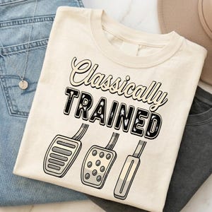 Może przedstawiać: Kremowy t-shirt z napisem "Classically Trained" pogrubioną, postarzaną czarną czcionką. Poniżej tekstu znajduje się graficzna ilustracja trzech pedałów samochodowych, przedstawiająca sprzęgło, hamulec i pedał gazu w manualnej skrzyni biegów.