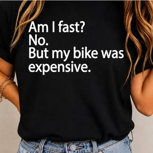 May include: A black short-sleeved t-shirt featuring white text printed on the front that reads: Am I fast? No. But my bike was expensive. The shirt is paired with blue denim jeans, and the design is centered on the chest area of the garment.