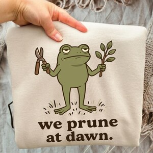 May include: A beige sweatshirt featuring a graphic of a green frog holding garden shears in one hand and a leafy branch in the other. Below the frog, the text reads, "we prune at dawn." The design is printed in dark brown ink on the fabric.