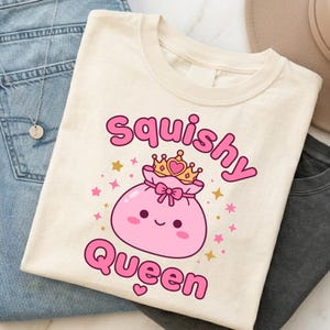 Może przedstawiać: Kremowy t-shirt z grafiką przedstawiającą okrągłą, różową, uśmiechniętą postać w złotej koronie. Napis "Squishy Queen" jest nadrukowany na różowo nad i pod postacią, w otoczeniu małych różowych i złotych gwiazdek.