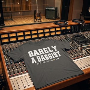 以下が含まれることがあります： ダークグレーの半袖Tシャツで、前面には「BARELY A BASSIST but i show up」という白いかすれた文字と、3つの金色の星、2つの灰色の星がプリントされています。プロ仕様のオーディオミキシングコンソールの上に平らに置かれています。