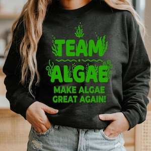 Könnte beinhalten: Ein schwarzes Langarm-Sweatshirt mit hellgrünem Text, der "TEAM ALGAE" und "MAKE ALGAE GREAT AGAIN!" lautet, sowie grafischen Darstellungen von Algen und Blasen, die den Schriftzug auf der Vorderseite des Kleidungsstücks umgeben.