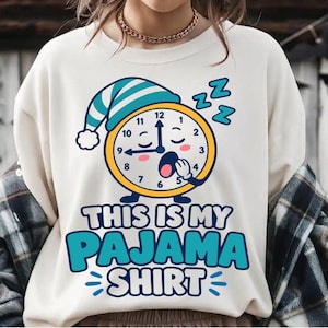May include: A cream-colored crewneck sweatshirt featuring a graphic of a yellow clock wearing a blue and white striped nightcap. The clock has a sleepy face and is yawning. Below the graphic, the text reads: This is my pajama shirt.