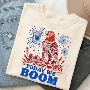 May include: Cream-colored t-shirt featuring a patriotic design. The graphic includes a red, white, and blue eagle wearing sunglasses, surrounded by fireworks and the text "TODAY WE BOOM" in blue. The shirt is paired with blue jeans and a necklace.