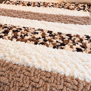 May include: A chunky knit throw blanket featuring horizontal stripes in cream, tan, and a speckled brown and black leopard print pattern. The thick, soft yarn creates a textured, looped appearance across the entire surface of the rectangular blanket.