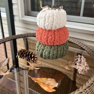 May include: A stacked decorative pumpkin ornament made of three crocheted yarn sections in white, orange, and olive green. The top section features a small brown stem with curled twine tendrils. The texture is soft, fuzzy, and ribbed.