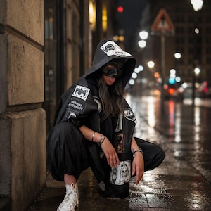 Puede incluir: Una capa negra con capucha que presenta varios parches gráficos blancos y texto, incluida la frase "NO BOSSES NO MANAGERS". La capa se lleva sobre pantalones negros y se combina con zapatillas blancas de caña alta. Un antifaz negro cubre el rostro.
