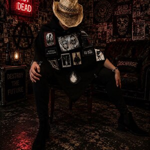 Puede incluir: Un sombrero de vaquero de paja usado con un poncho negro que presenta varios parches rectangulares con ilustraciones gráficas y textos que incluyen "NO BOSSES NO MANAGERS" y "SEE INTO THE DARK," junto con pantalones y botas negras.