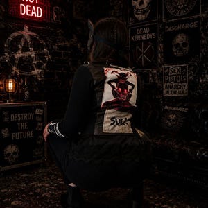Peut inclure: Une veste noire avec un grand écusson rectangulaire dans le dos représentant une créature aux yeux rouges et le texte SUCK. La veste présente des manches longues, une coupe ajustée et une bordure en dentelle noire le long de l'ourlet inférieur.