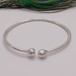 May include: A silver-colored open bangle bracelet with a smooth, rounded design. The bracelet features a ball-shaped end on each side. A peacock feather is in the background.