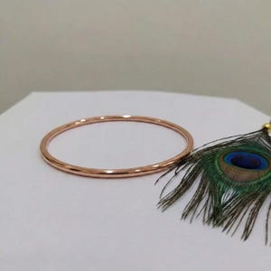 May include: A thin, circular rose gold bangle bracelet with a smooth, polished metallic finish. The bangle is positioned next to a single peacock feather featuring vibrant blue, green, and brown eye-patterned plumage on a plain white surface.