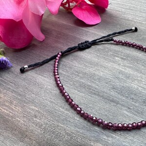 May include: A delicate bracelet featuring small, faceted deep purple beads strung on a thin black cord. The bracelet includes an adjustable sliding knot closure with two small silver-toned metal beads at the ends of the cord for a secure fit.