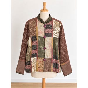 May include: A long-sleeved jacket featuring a patchwork design with various fabric textures and patterns in shades of brown, olive green, and cream. The jacket has a mandarin collar, four purple buttons down the front, and solid brown sleeves.