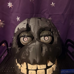 May include: A dark gray, weathered-looking mask featuring large, round, light-colored eyes with dark pupils and a wide, toothy grin. The mask has a distressed, damaged texture with jagged edges around the eye sockets and mouth area.
