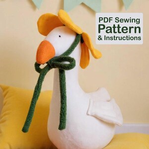 May include: A plush white goose with an orange beak and feet, wearing a yellow flower crown and a green scarf. The goose is sitting on a yellow pillow. The image includes text that says "PDF Sewing Pattern & Instructions."