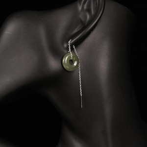 May include: A single earring featuring a circular, olive-green jade pendant with a hole in the center. The pendant hangs from a delicate silver chain. The earring is displayed on a black mannequin head against a black background.
