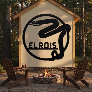 Pode incluir: Uma grande placa de metal preta com o texto "EL ROIS" e um desenho de cobra é montada em uma estrutura de madeira. Duas cadeiras de madeira em frente a uma fogueira com fogo. Duas garrafas em uma pequena mesa.