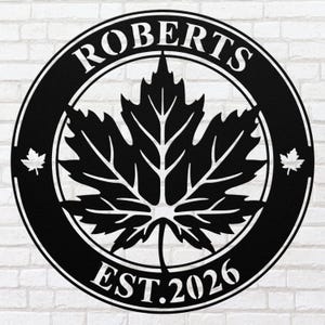 May include: Black metal wall art with a maple leaf design. The circular piece includes the name "ROBERTS" and the year "EST. 2026". Two small maple leaf accents are on either side of the main leaf.