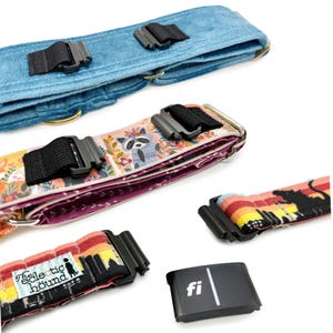 May include: A collection of dog collars in various patterns and colors. One collar is a light blue velvet, another features a raccoon print, and others display sunset and cityscape designs. Each collar has a black buckle and a gold-colored metal ring.