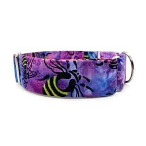 May include: A dog collar with a purple, blue, and black bee pattern. The collar has a metal D-ring and a silver-colored buckle. The bee design features black outlines and yellow stripes on the bees.