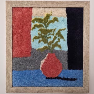 May include: A framed textile art piece featuring a red vase with a green plant against a backdrop of blue, red, gray, and black textured panels. The artwork is encased in a light brown wooden frame, creating a modern, abstract still life.