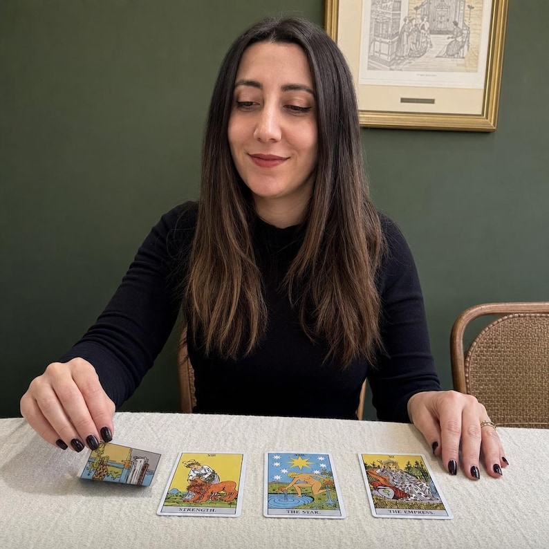 May include: A set of four tarot cards laid out on a light-coloured surface. The cards include Strength, The Star, and The Empress, with one card being held upright. Each card features detailed illustrations with vibrant colours and clear black text labels.