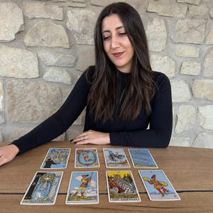 May include: A set of eight tarot cards arranged in two rows on a wooden surface. The cards feature various illustrations including The High Priestess, The Empress, Knight of Cups, and other figures, with text labels visible on several of the cards.