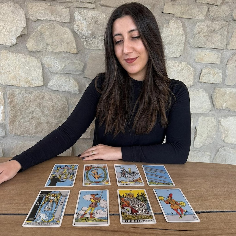 May include: A set of eight tarot cards arranged in two rows on a wooden surface. The cards feature various illustrations including The High Priestess, The Empress, Knight of Cups, and other figures, with text labels visible on several of the cards.