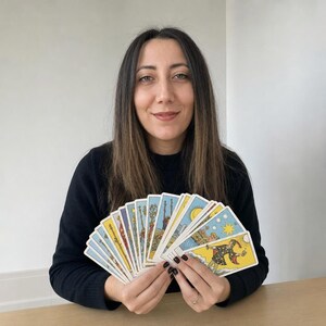 May include: A fan of tarot cards held in two hands, featuring colorful illustrations including the Star and the Fool cards. The cards display detailed artwork with yellow, blue, and red tones, showcasing various symbolic figures and celestial imagery.