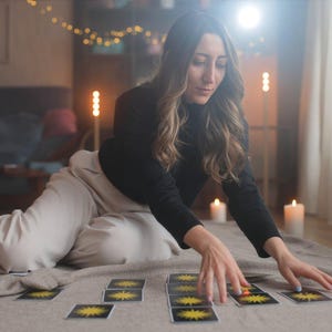 May include: A person is arranging tarot cards with a yellow sun design on a neutral-colored surface. The person is wearing a black long-sleeved shirt and light-colored pants. Lit candles and string lights create a warm ambiance.
