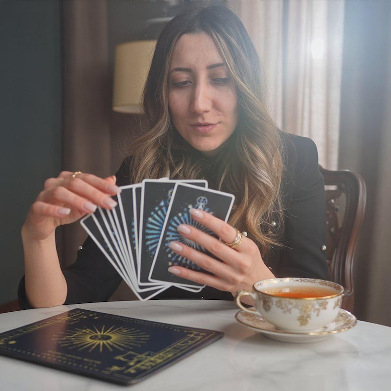 May include: A woman holding a deck of tarot cards with a blue and white design. A gold-rimmed teacup and saucer sit on a white marble table. A dark blue mat with a gold sunburst design is also on the table.