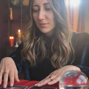 May include: A woman with long, wavy hair sits at a red table, surrounded by candles and a crystal ball. Tarot cards with yellow sun designs are laid out. The scene suggests a mystical or fortune-telling theme.