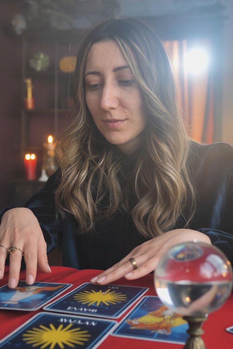 May include: A woman with long, wavy hair sits at a red table, surrounded by candles and a crystal ball. Tarot cards with yellow sun designs are laid out. The scene suggests a mystical or fortune-telling theme.