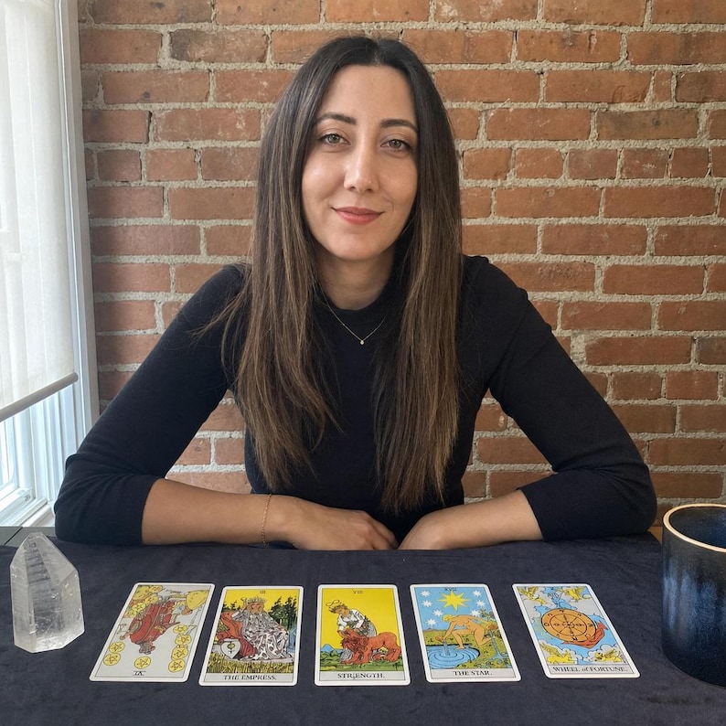 May include: A set of five tarot cards laid out on a dark surface, including The Empress, Strength, The Star, and Wheel of Fortune. To the left is a clear quartz crystal point, and to the right is a dark blue ceramic cup. The cards feature colorful illustrations.
