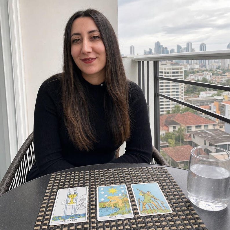 May include: Three tarot cards laid out on a table, including the Ace of Cups, The Star, and the Seven of Wands. The cards feature detailed illustrations with blue, yellow, and green tones, positioned next to a clear glass of water on a mat.