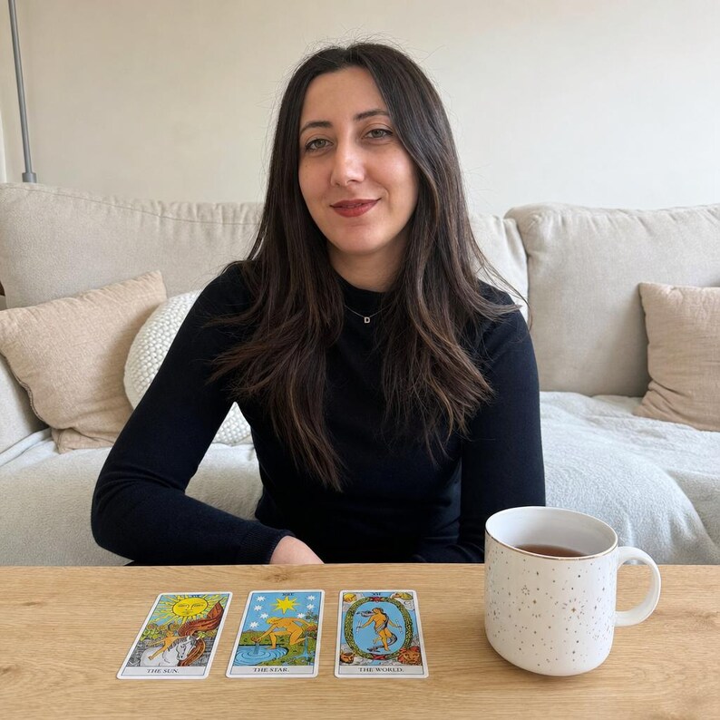 May include: Three tarot cards laid out on a wooden surface, featuring The Sun, The Star, and The World, positioned next to a white ceramic mug with a gold speckled pattern. The cards display colorful illustrations and their respective titles.