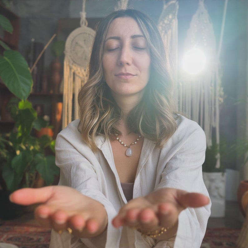 May include: A person with closed eyes, wearing a white shirt and a necklace with a pendant, extends open hands towards the viewer. The background includes plants, macrame wall hangings, and a soft, bright light.
