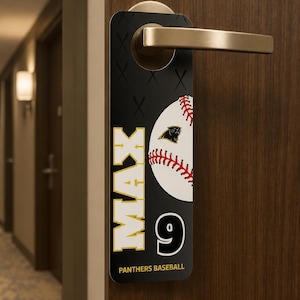 Op de afbeelding: Zwarte deurhanger met een honkbalontwerp. De tekst "MAX 9" is in wit en geel. De honkbal heeft rode stiksels en een teamlogo. De woorden "Panthers Baseball" staan onderaan.