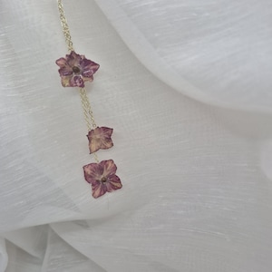 Peut inclure: Un collier à chaîne dorée délicat orné de trois pétales de fleurs d'hortensia violettes pressées fixés à intervalles. Les pétales présentent des nervures naturelles et des nuances variées de violet et de mauve, suspendus verticalement.