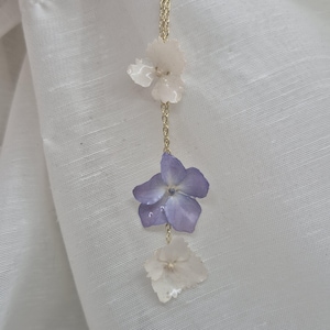Peut inclure: Un collier à chaîne dorée orné de trois breloques en forme de fleurs disposées verticalement. Les fleurs du haut et du bas sont blanches, tandis que la fleur centrale est d'un violet lavande doux. Chaque fleur a cinq pétales à la finition lisse.
