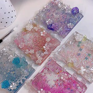 May include: A collection of five square resin coasters. Each coaster features a floral design with glitter and small embellishments in shades of pink, blue, purple, and clear. The coasters have a decorative edge.