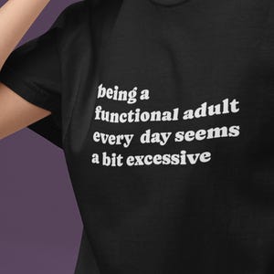 Könnte beinhalten: Schwarzes T-Shirt mit dem weißen Text "being a functional adult every day seems a bit excessive". Das Shirt hat einen Rundhalsausschnitt und kurze Ärmel. Das Bild ist eine Nahaufnahme des Shirts.