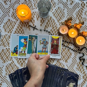 Puede incluir: Una configuraci&oacute;n de lectura de cartas del tarot con velas encendidas, un rosario y una baraja de cartas. Las cartas est&aacute;n dispuestas sobre un mantel de encaje blanco, con una mano sosteniendo las cartas.