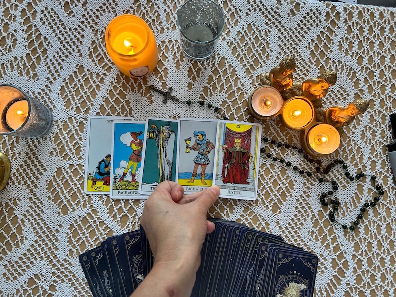 Puede incluir: Una configuraci&oacute;n de lectura de cartas del tarot con velas encendidas, un rosario y una baraja de cartas. Las cartas est&aacute;n dispuestas sobre un mantel de encaje blanco, con una mano sosteniendo las cartas.